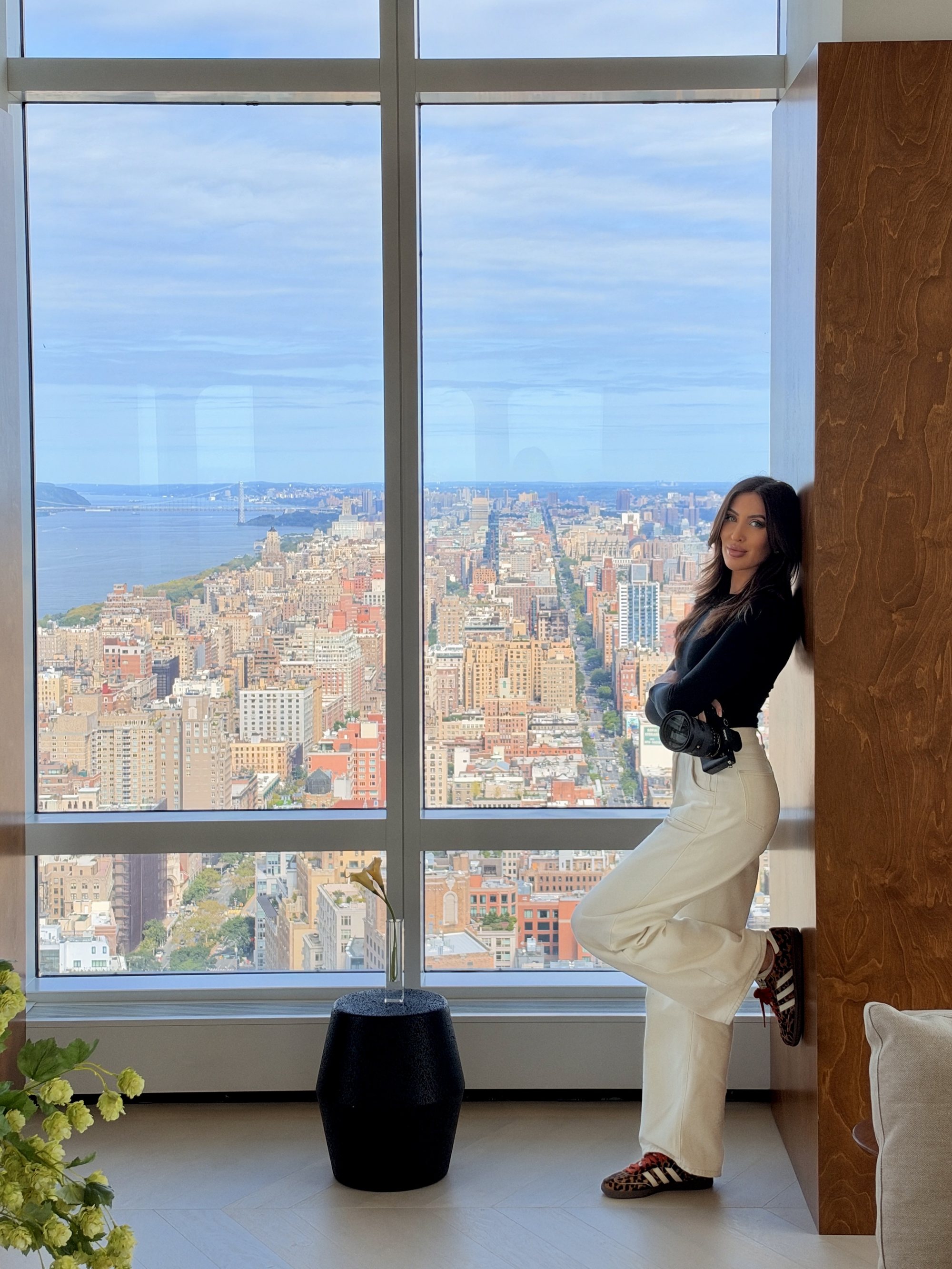 Sarah Cotty leaning against a wood wall, camera in her arms, looking toward the camera with the city behind her.