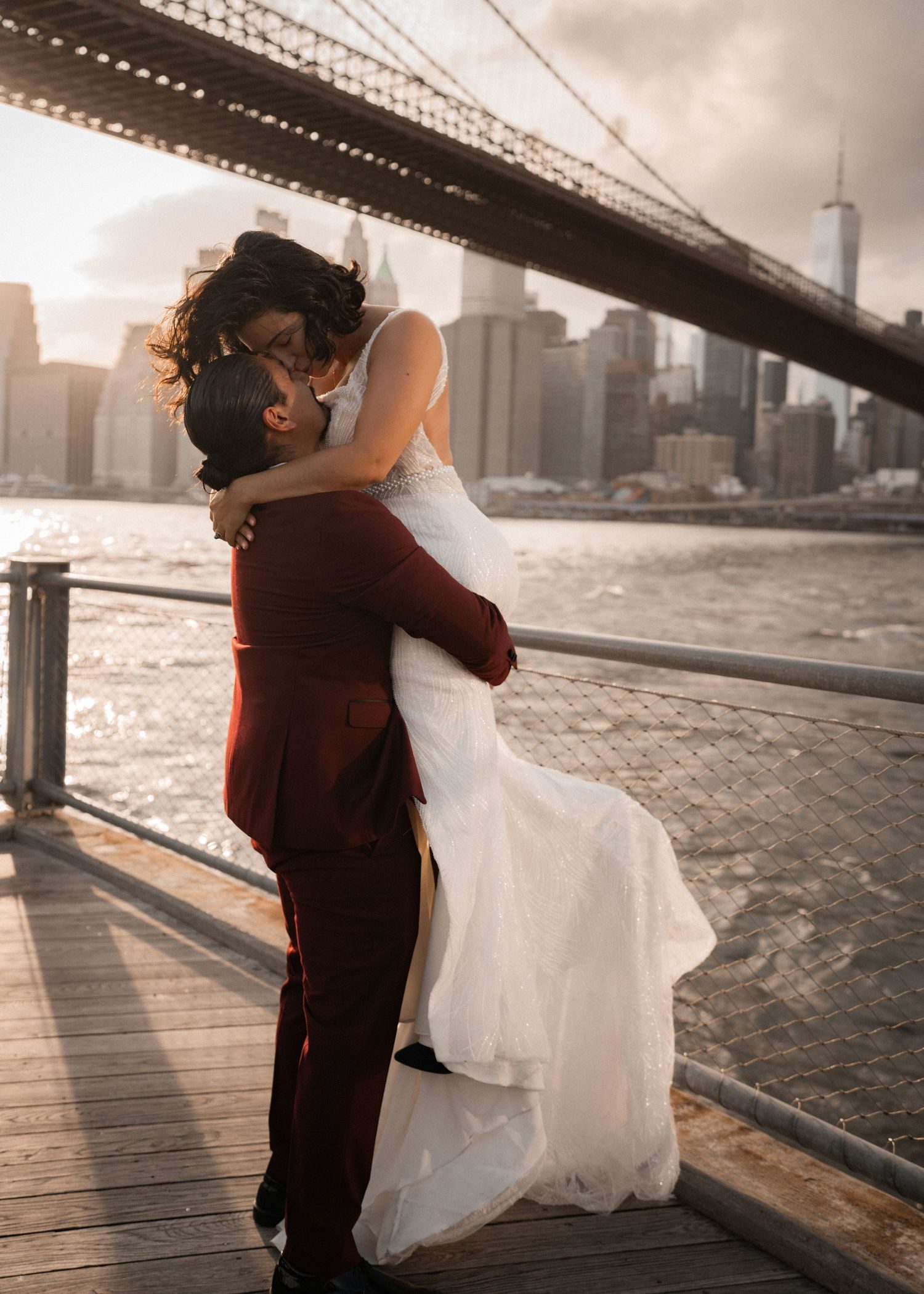 A couple on a waterfront, him lifting her, a bridge and the skyline behind them.