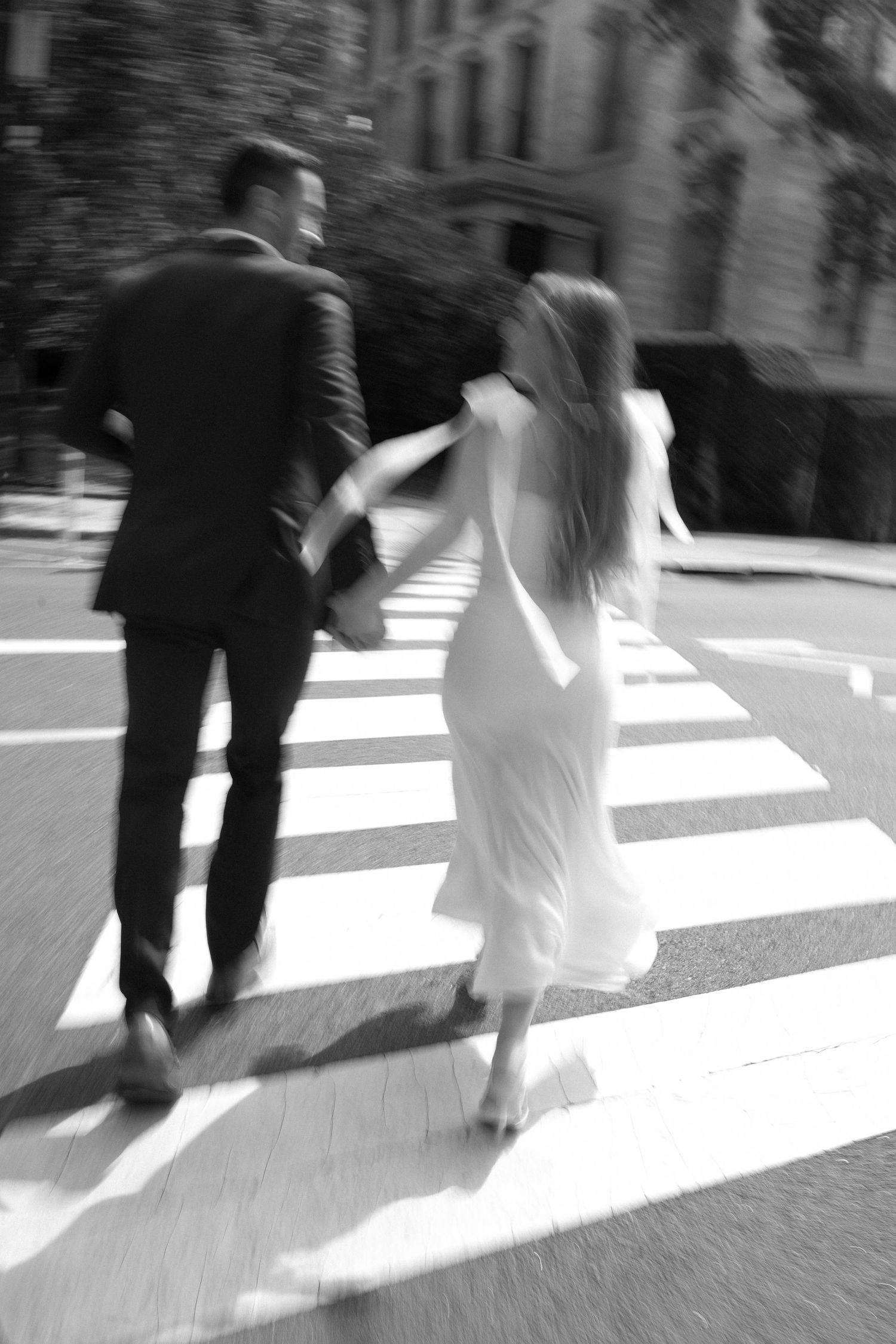 A couple in black and white crossing a city street, her dress caught in the wind.