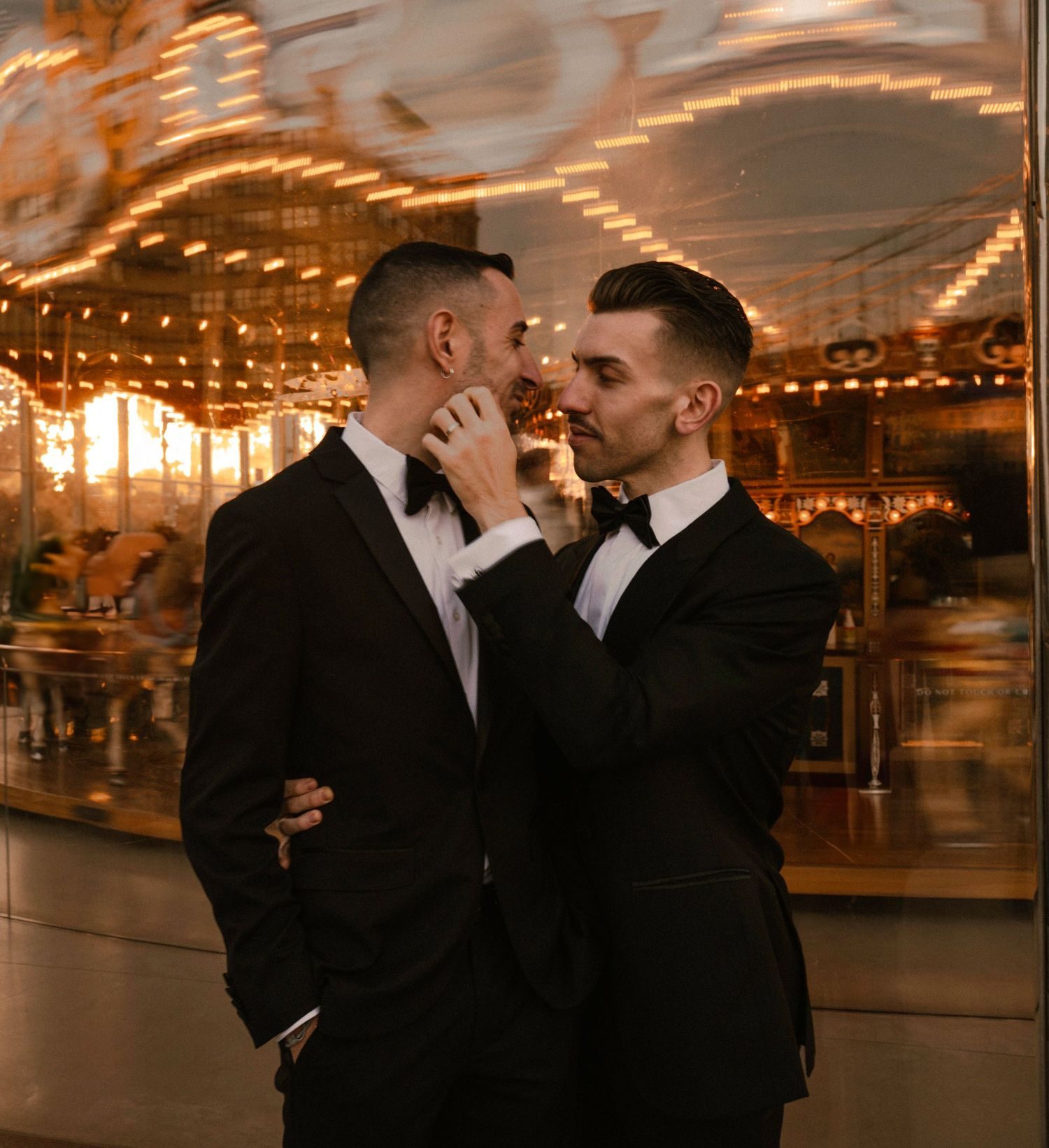 Two grooms in black tuxedos in front of a lit carousel.