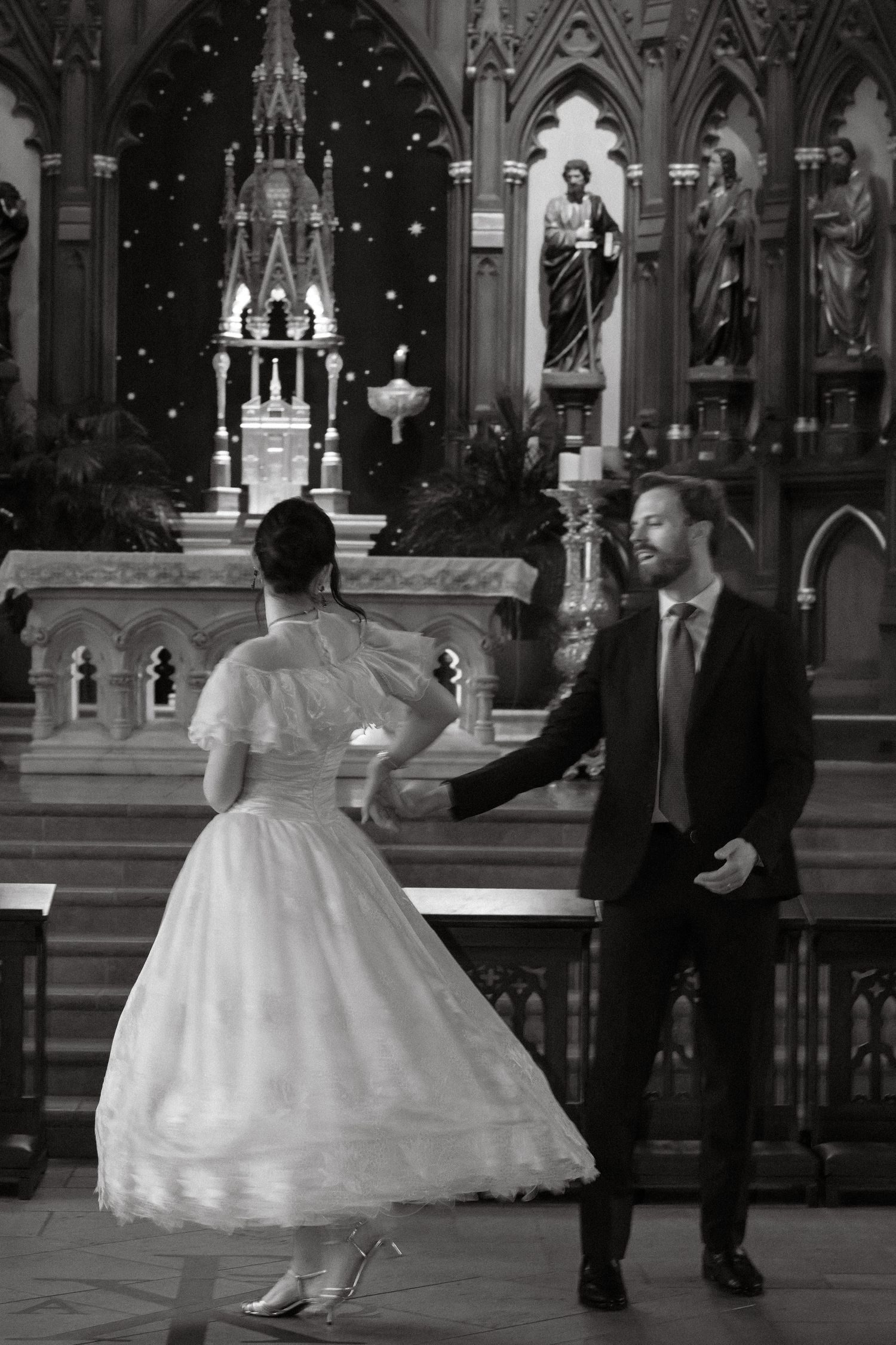 A bride spinning in a full skirt inside a church, the groom beside her, in black and white.