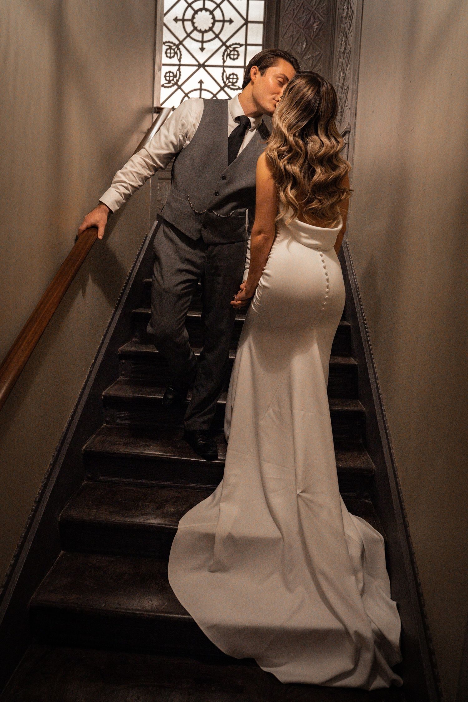 A bride and groom kissing on a staircase, her gown spilling down the steps.